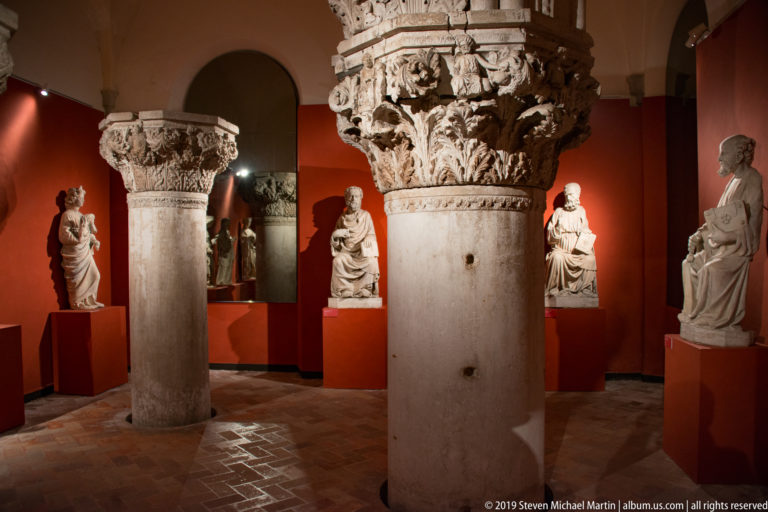Large carved stone columns and capitals surrounded by pale sculptures on red pedestals in a museum gallery.