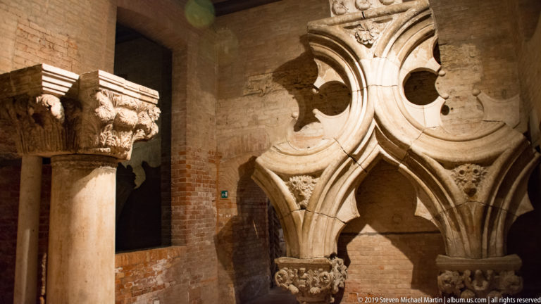 Carved stone capitals and a large Gothic tracery fragment displayed against an exposed brick wall.
