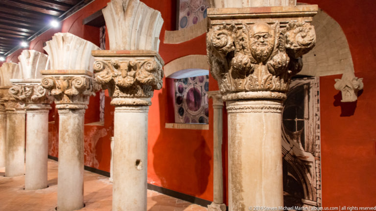 Stone columns with varied carved capitals featuring foliage, faces, and figures in a red-walled museum gallery.