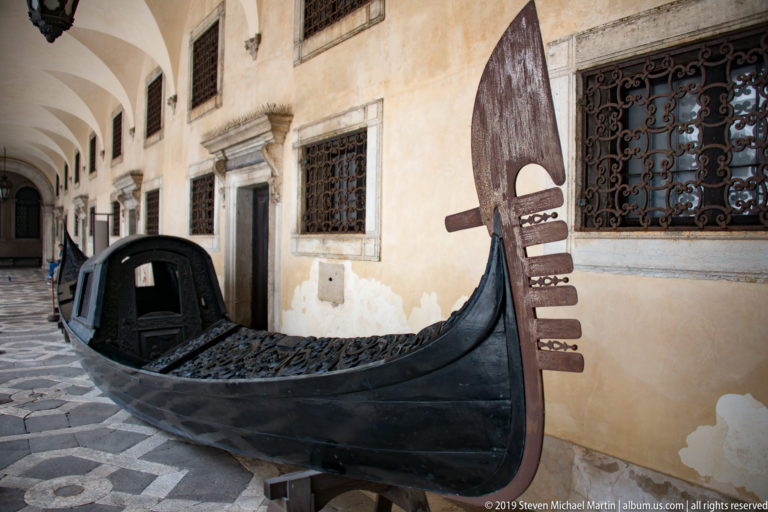Historic black Venetian gondola displayed indoors beside a plaster wall with iron-grilled windows.