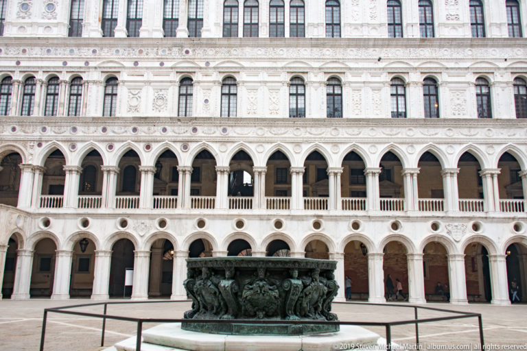Bronze wellhead centered before three tiers of pale arcades in the Doge’s Palace courtyard.