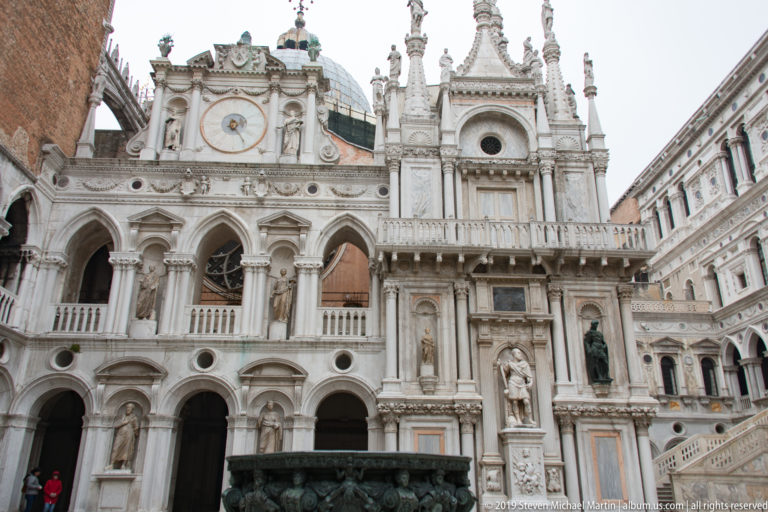 Bronze wellhead before the ornate courtyard façade of the Doge’s Palace, with clock, pinnacles, arcades, statues, and staircase.