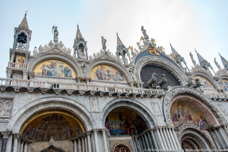 Oblique low-angle view of St. Mark’s Basilica with mosaic portals, central window, statues, pinnacles, and carved arches.