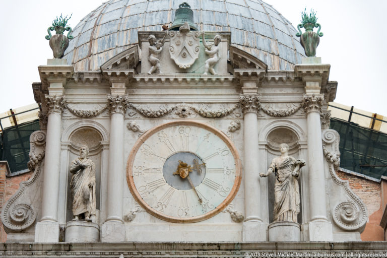 Large circular clock set in an ornate stone façade with flanking statues, scrollwork, and a metal-clad roof.