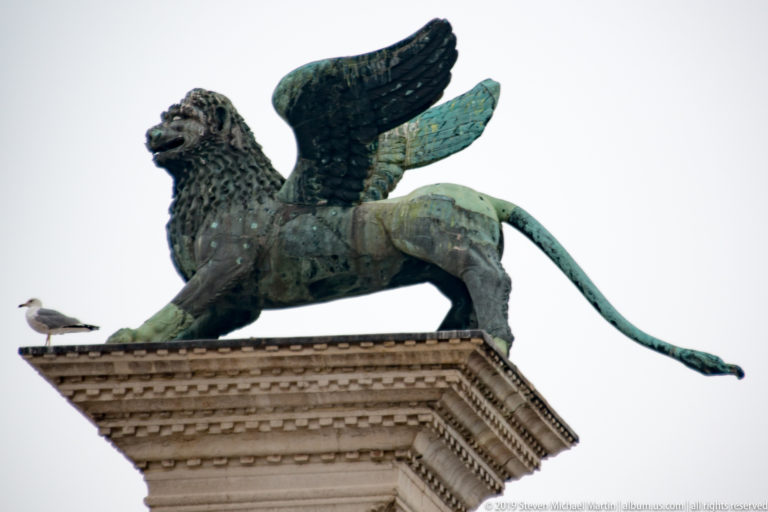 Bronze winged Lion of St. Mark standing atop a stone column capital with a gull perched nearby.