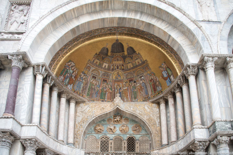 Gold mosaic lunette above a pointed sculpted opening, framed by clustered marble columns and arches on St. Mark’s Basilica.
