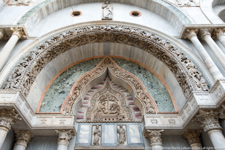 Pointed carved arch with relief figures, foliage, colored stone insets, columns, and capitals on St. Mark’s Basilica.