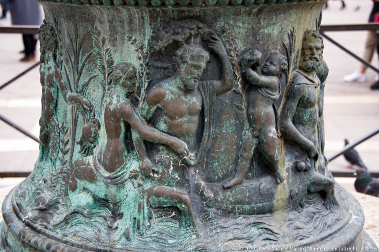 Patinated bronze relief with a muscular nude figure, smaller attendant, and foliage on a historic wellhead.