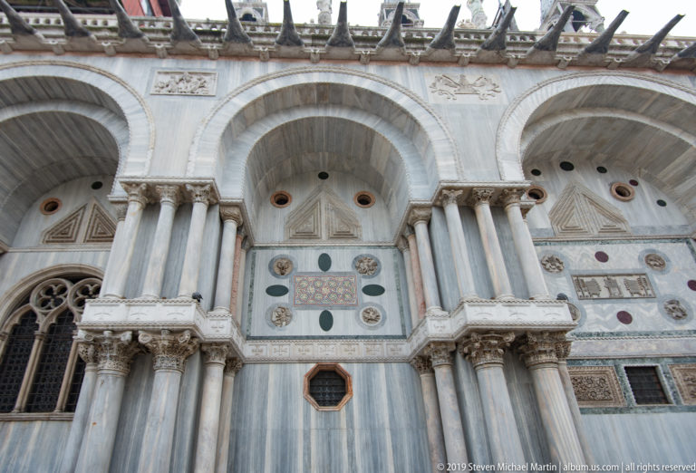 Three marble arches on clustered columns above geometric panels, colored stone disks, reliefs, and small windows on St. Mark’s Basilica.