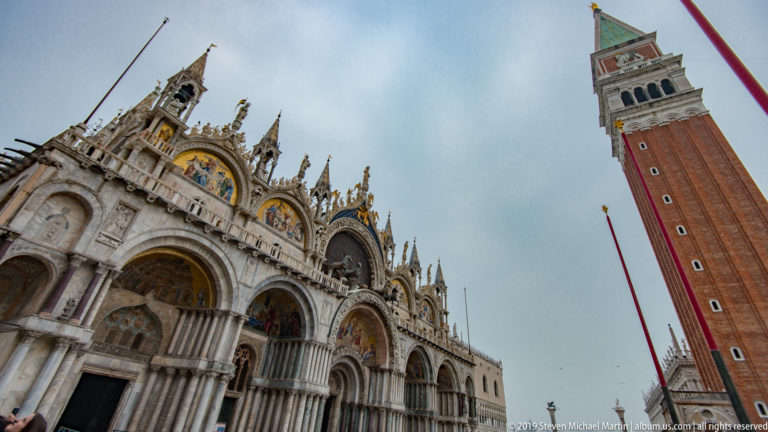 Low-angle view of St. Mark’s Basilica façade at left and the red-brick Campanile rising at right under a cloudy sky.