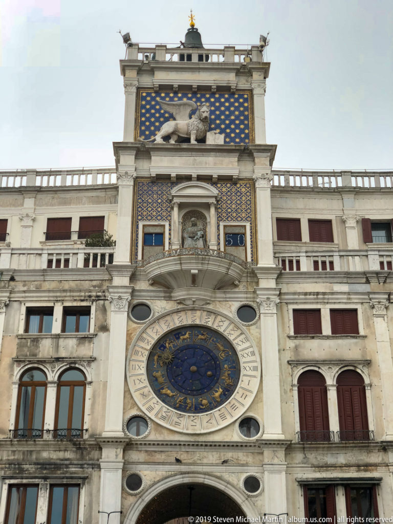 St. Mark’s Clocktower façade with blue astronomical clock, winged lion relief, arched windows, and sculptural upper tiers.