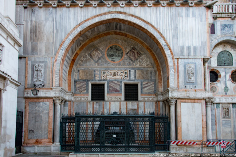 Side arch of St. Mark’s Basilica with striped stone voussoirs, marble panels, reliefs, and a dark metal gate.