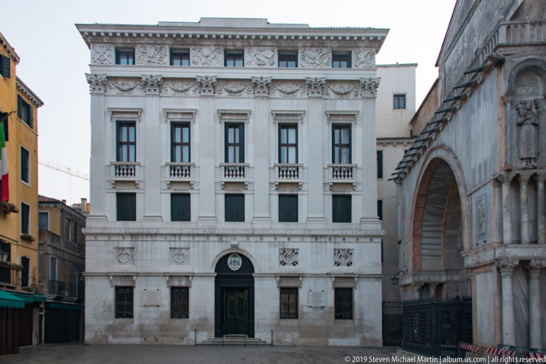 Symmetrical pale palace façade with rows of windows and central doorway beside the marble exterior of St. Mark’s Basilica.