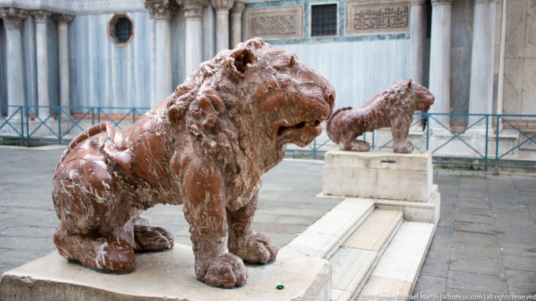 Two weathered reddish stone lion sculptures on plinths beside the pale marble exterior of St. Mark’s Basilica.