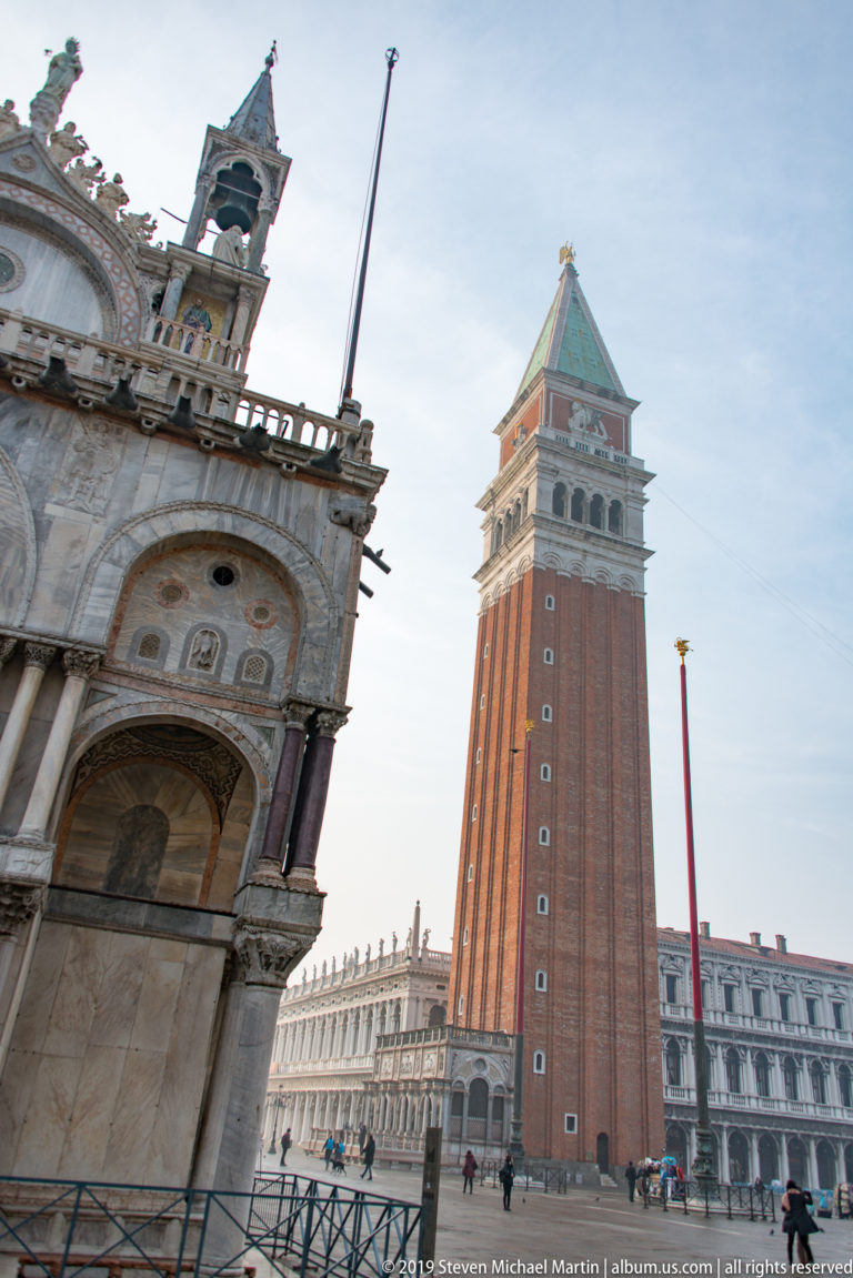 Red-brick St. Mark’s Campanile rising beside the carved marble corner of St. Mark’s Basilica from a low angled viewpoint.