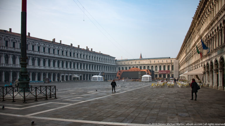 Broad Piazza San Marco with patterned paving, long arcaded façades, sparse pedestrians, and buildings closing the far end.