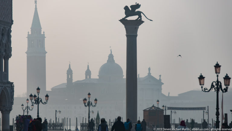 Winged lion statue atop a tall column silhouetted against hazy domes, a distant campanile, lamps, and pedestrians.