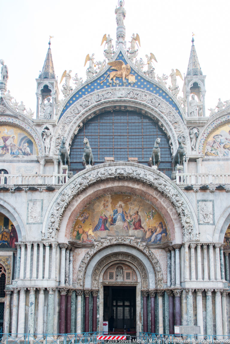 Central entrance of St. Mark’s Basilica beneath carved arches, mosaic lunettes, upper window, and gilded roofline.