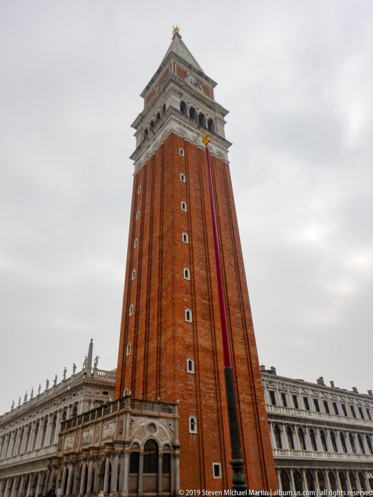 Tall red-brick St. Mark’s Campanile with pale belfry and green roof rising above the marble Loggetta and arcades.