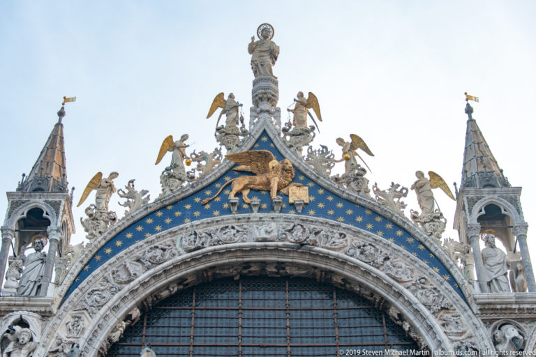 Upper arch of St. Mark’s Basilica with gilded figures, blue mosaic field, carved stonework, and pinnacled niches.