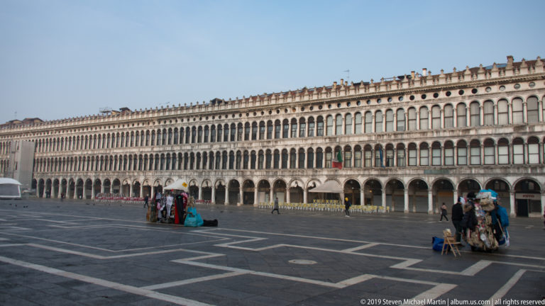 Long arcaded Procuratie façade bordering the patterned paving of Piazza San Marco with a few pedestrians and vendors.