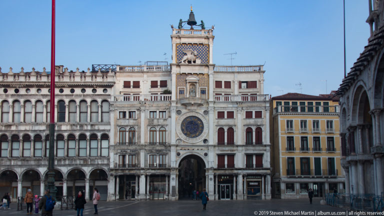 St. Mark’s Clock Tower centered among arcaded façades, with pedestrians crossing the paved Piazza San Marco.
