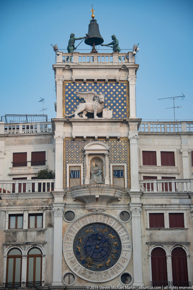 Astronomical clock face beneath the winged lion panel and rooftop bell of St. Mark’s Clock Tower.