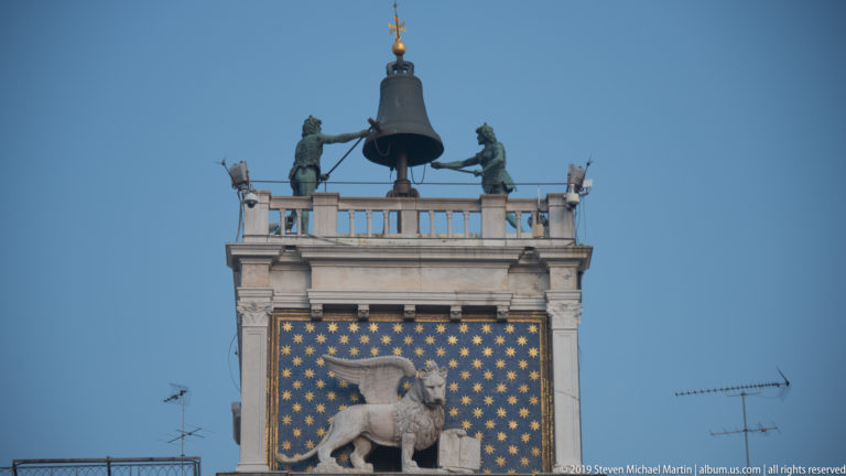 Winged lion mosaic panel below the rooftop bell and two bronze figures on St. Mark’s Clock Tower.