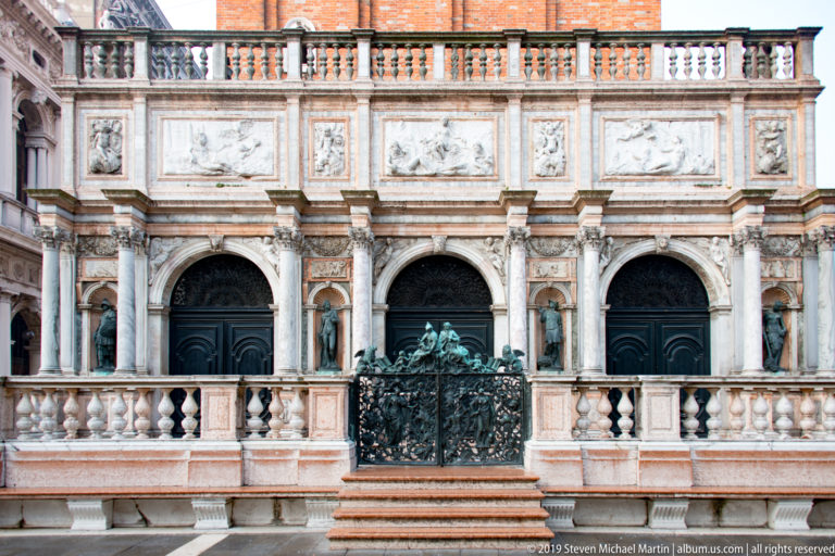 Ornate marble Loggetta façade with paired columns, relief panels, bronze statues, balustrade, and central metal gate.