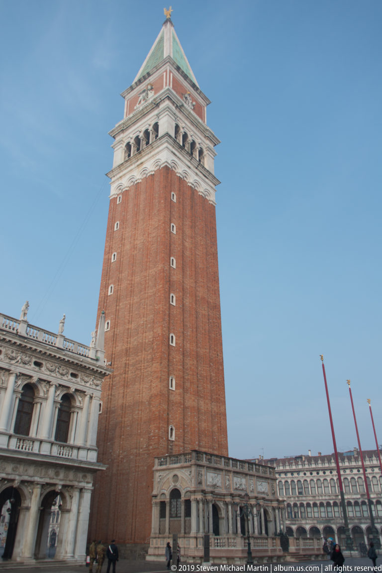 Brick campanile rising above the marble Loggetta and arcaded buildings under a clear blue sky.