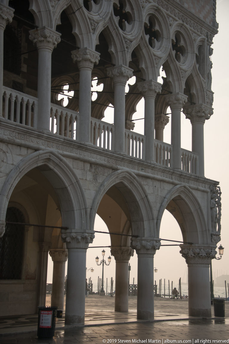 Backlit corner of the Doge’s Palace with lower arches, upper quatrefoil arcade, slender columns, and lagoon beyond.
