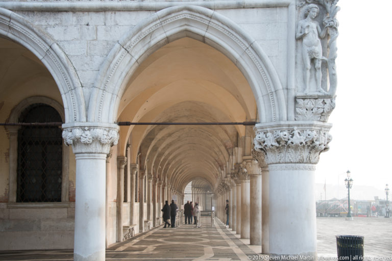 Long Doge’s Palace arcade with pointed arches, carved capitals, patterned pavement, and a few pedestrians receding into the distance.