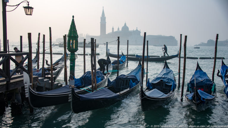 Clustered gondolas at a waterfront landing with wooden mooring poles, green lantern, and hazy San Giorgio Maggiore beyond.