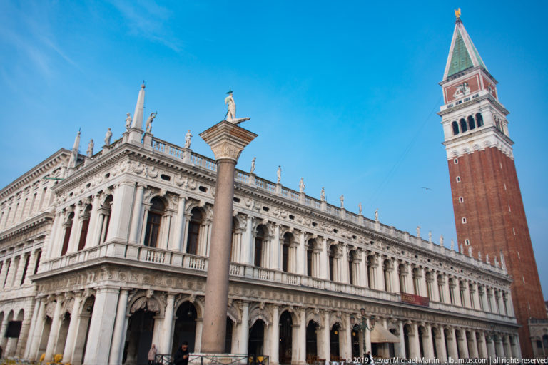 Marciana Library arcades beside St. Mark’s Campanile, with a monumental Piazzetta column and rooftop statues in the foreground.