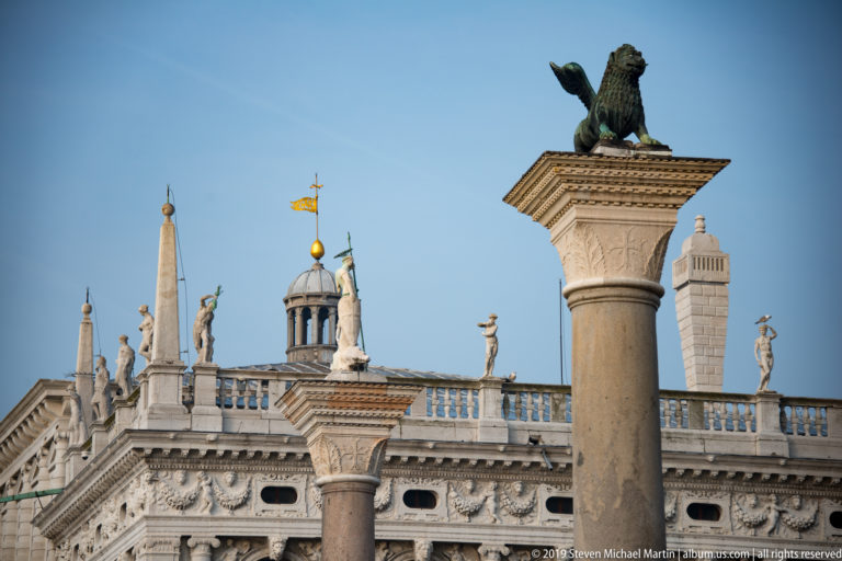 Winged lion statue atop a tall stone column with the statue-lined roof of the Marciana Library behind it.