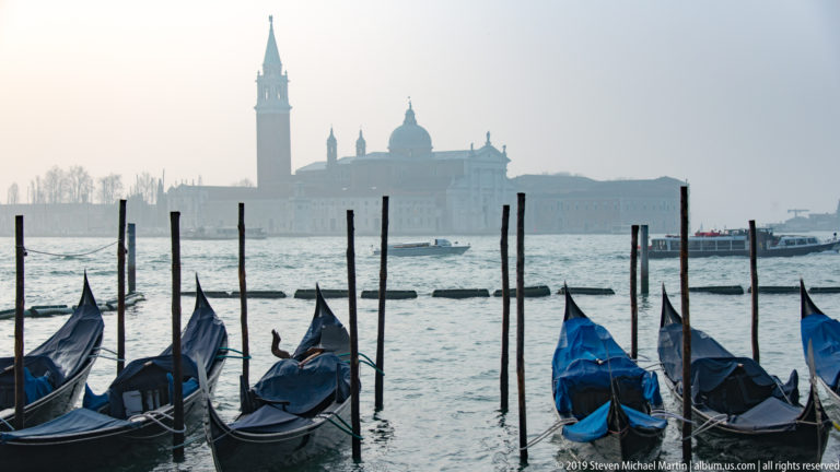 Moored gondolas and wooden poles on the lagoon with hazy San Giorgio Maggiore church and campanile across the water.