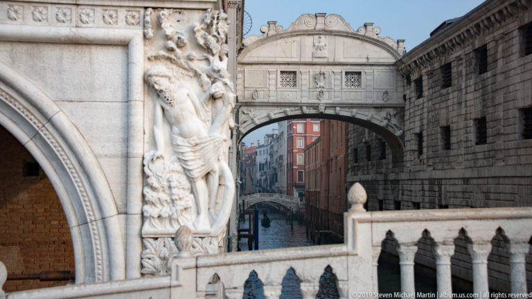 Carved stone figure and pointed arch on the Doge’s Palace foreground the Bridge of Sighs over the canal behind.