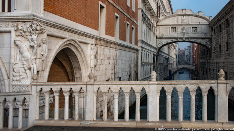 Bridge of Sighs beyond a carved marble balustrade, with sculpted palace portal at left and canal extending behind it.