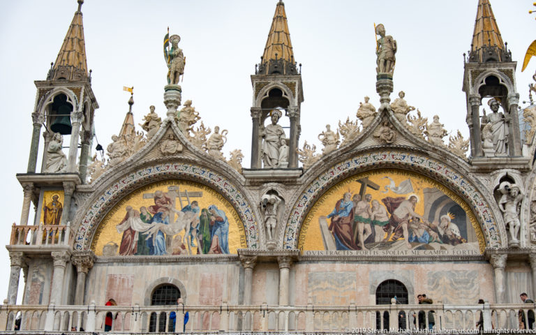 Two gold mosaic lunettes beneath sculptural bell turrets, pinnacles, and statues on St. Mark’s Basilica.