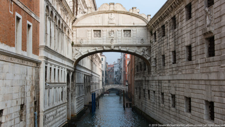 Bridge of Sighs over the narrow Rio di Palazzo, with stone palace walls, canal water, and another bridge in the distance.