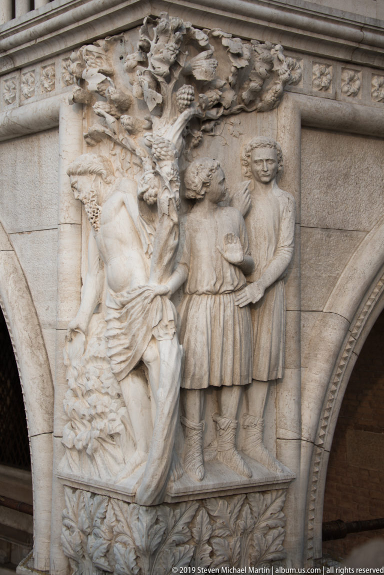 Carved stone relief with a dynamic central figure, standing attendants, foliage, and adjacent pointed arch on the Doge’s Palace.