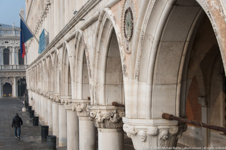 Long perspective down the Doge’s Palace arcade with pointed arches, carved capitals, columns, and a pedestrian below.