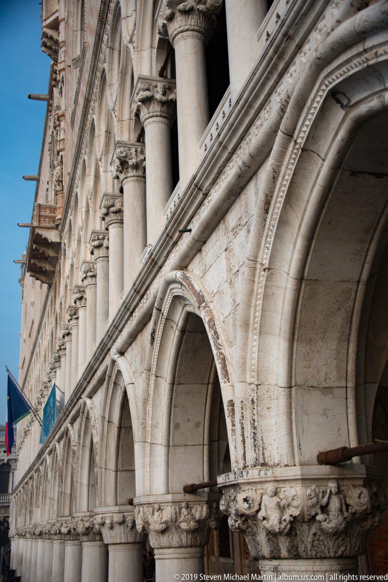 Diagonal close view along the Doge’s Palace arcade, showing pointed stone arches, columns, carved capitals, and Gothic detailing.