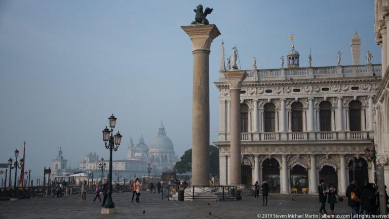 Piazzetta San Marco with monumental column, Marciana Library façade, pedestrians, and hazy San Giorgio Maggiore across the lagoon.