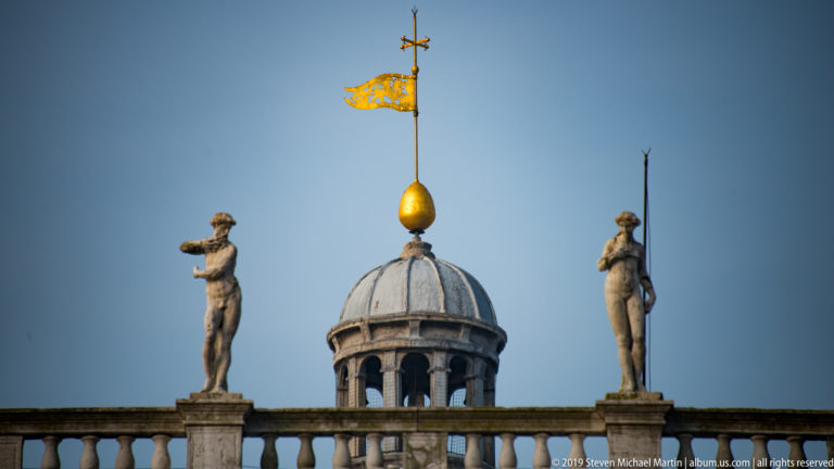 Lead-covered rooftop cupola with gilded orb and banner, flanked by two standing stone statues on a balustrade.
