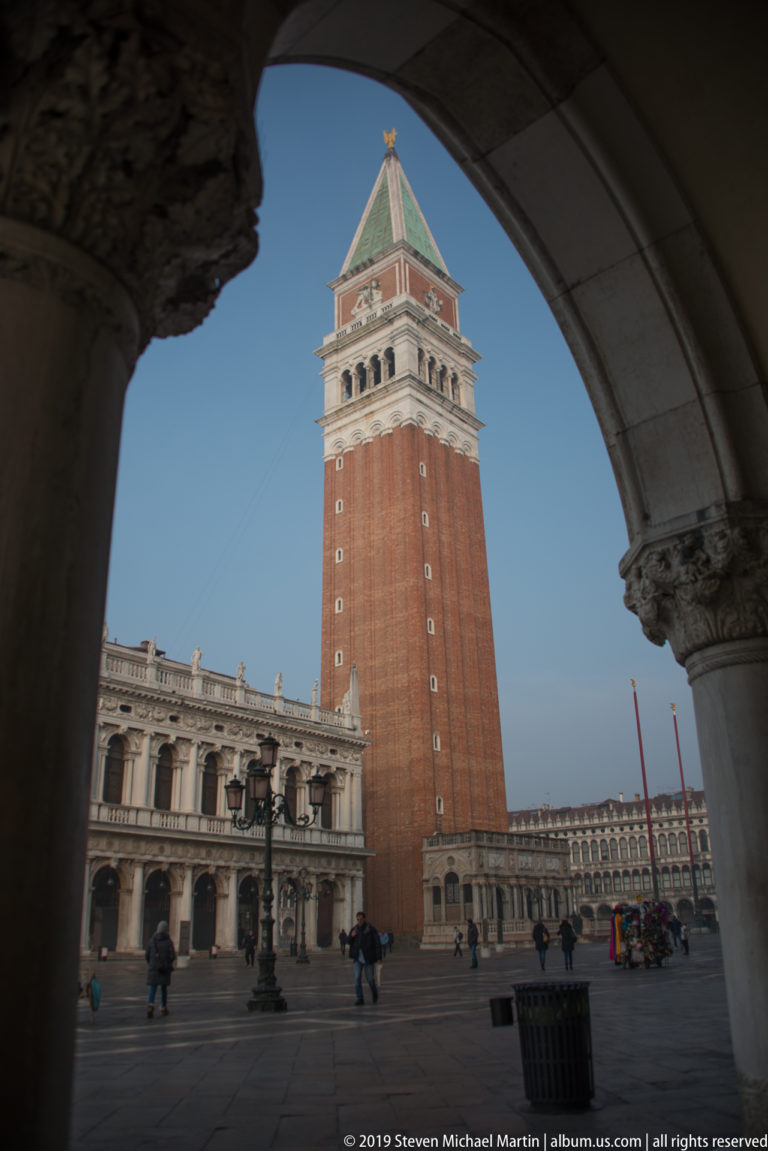 St. Mark’s Campanile framed through a shadowed stone arch, with arcaded square façades and pedestrians below.