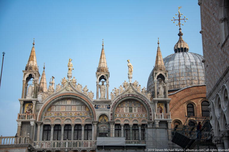 Upper façade of St. Mark’s Basilica with pinnacles, statues, arched mosaic panels, and a lead-covered dome.