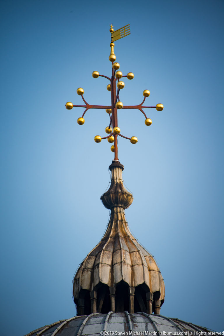 Gilded branching rooftop finial and weather vane mounted on a lead-covered cupola against a clear blue sky.
