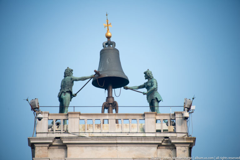 Large bell flanked by two green-patinated bronze striking figures atop St. Mark’s Clocktower against a blue sky.