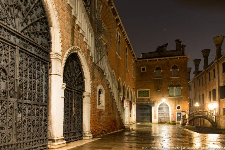 Ornate Gothic doorway and iron gate on a brick façade under nighttime illumination in Venice.
